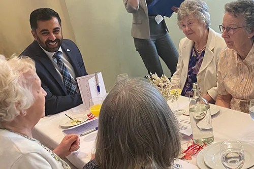 First Minister Humza Yousaf smiling and engaging in lively conversation with a group of NHS Retirement Fellowship members at an informal gathering. The group appears cheerful and relaxed, capturing a moment of warmth and connection.