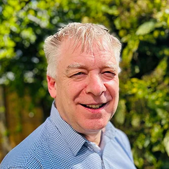 Head-and-shoulders portrait of Rod How, Chief Operating Officer for the NHS Retirement Fellowship. He is smiling and dressed in a checked blue shirt, standing outside against a blurred green background.