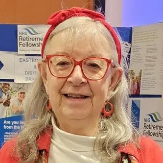 Head-and-shoulders portrait of Jane Dubery, a representative of the Members' Council for the NHS Retirement Fellowship. She is smiling, wears red glasses, a red headband and a red sweater, and standing against a busy noticeboard.